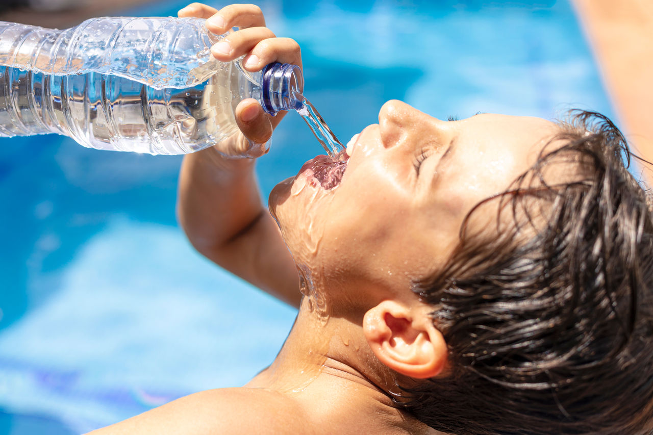 Boy drinking water from a plastic bottle in in the pool on a hot summer day. Young man drinking a lot of water after sports to avoid heat stroke. Child drinking plenty of water after physical exercise