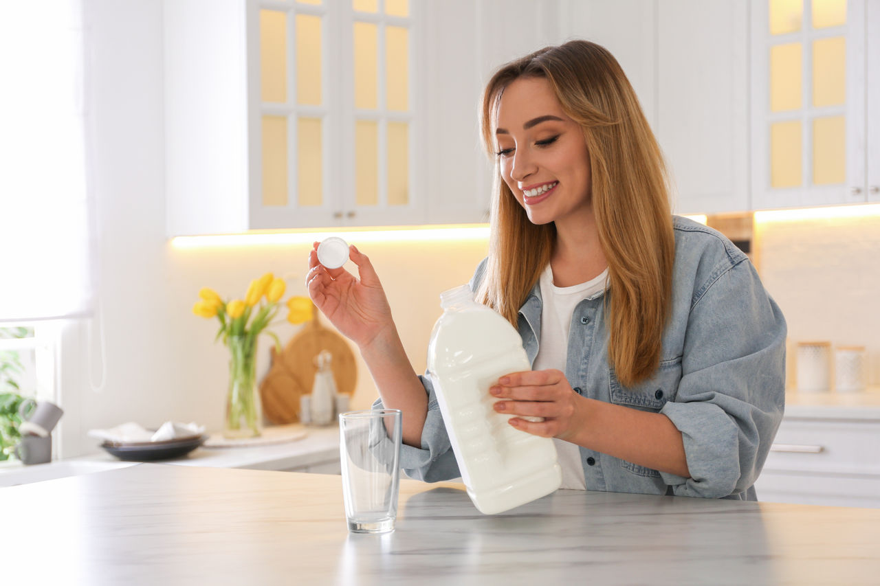Young woman pouring milk from gallon bottle into glass at white marble table in kitchen; Shutterstock ID 2208427439; purchase_order: POL website_OmyaPET; job: ; client: ; other: 